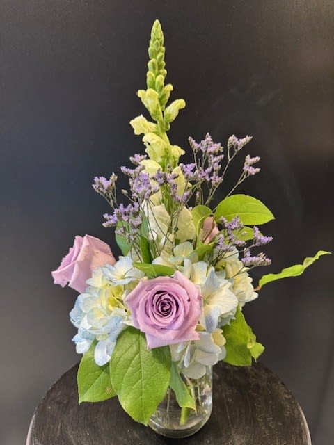 #A087 - Pastel Passion - Soft lavender roses tucked among pale blue and white hydrangeas create a calming, pastel statement - perfect as a birthday bouquet, Mother's Day surprise, or a gentle get well arrangement. This flower arrangement for Spring or Easter (yet lovely any time of year) pairs romantic lavender and soft pink tones with touches of green and blue to convey tenderness and calm; it's the kind of gift that makes the person you're thinking of feel comforted and seen. Compact and fresh, the design features hydrangeas, roses, a single tall white snapdragon spike for subtle height, and fine sprays of small purple filler flowers for a delicate, airy texture. Arranged in a clear glass vase, it fits neatly on a desk, bedside table, or small dining surface-ideal for housewarming, graduation, anniversary, new baby celebrations, or simply saying "thank you" or "thinking of you." Lightly mist blooms daily and keep vase water fresh to extend enjoyment.