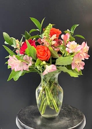 #A092 - Soft Romance - Bright orange roses take center stage in this loose, garden-style flower arrangement, making it an unforgettable birthday bouquet or anniversary surprise that reads as joyful and heartfelt. Designed for occasions from Mother's Day and Valentine's Day to a simple "thinking of you," the vivid mix of pink alstroemeria and red accents creates warm, affectionate energy while the tall mixed greens add fresh vertical movement and a hint of springtime optimism.   This mixed arrangement is presented in a clear glass vase filled with water for a casual, just-cut-from-the-garden look. It pairs soft carnation texture with the graceful spikes of snapdragons and abundant foliage, offering a lively palette of pink, orange, green, and red that brightens any room. The airy shape and varied heights make it a versatile choice for housewarmings, get well wishes, graduations, or to celebrate a new baby. Perfect for seasonal moments like Easter or as a thoughtful "I'm sorry" or thank-you gift, the bouquet is made to make the recipient feel genuinely seen and cheered. Vase-ready for immediate display, it's low-maintenance-refresh the water every few days and trim stems as needed-to keep the colors vivid through spring, summer, or into autumn and winter celebrations.