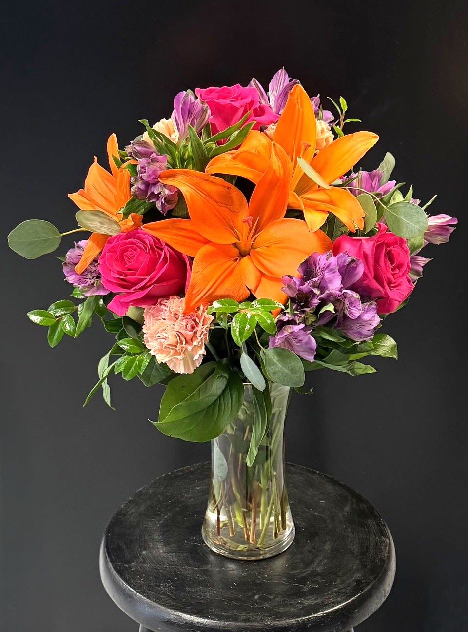 #A017 - Stunning Blossoms - Vivid orange Asiatic lilies take center stage in this lively flower arrangement, paired with hot pink roses and clusters of purple alstroemeria to create a saturated, celebratory bouquet perfect for a birthday bouquet, anniversary roses moment, or a Mother's Day gift. Soft peach carnations weave between the brighter blooms, giving a gentle color transition that keeps the overall look energetic without feeling overwhelming-an arrangement that reads as joyful, bold, and unmistakably festive whether you're marking a graduation, saying thank you, or sending love and romance.  Arranged upright in a clear glass vase, the gathered stems and clean lines give this design a classic everyday presentation that suits housewarmings, get well wishes, retirement parties, or a bright "just because" surprise. Size and structure make it equally at home on a dining table or office desk; the mix of textures-velvety roses, ruffled carnations, crisp lilies, and starry alstroemeria-creates movement and scent without excess. Seasonal enough for Easter or spring gifting yet timeless for year-round occasions like Thanksgiving, Valentine's Day, or thinking of you, this bouquet sends a clear message: the person you're thinking of will feel seen, celebrated, and genuinely uplifted.
