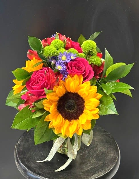#A098 - Sunflower Heaven - A bold yellow sunflower takes center stage in this cheerful flower arrangement, making it a joyful choice for a birthday bouquet, Mother's Day surprise, or just-because pick-me-up. Sunflower Heaven blends the sunny confidence of sunflowers with romantic hot pink roses and vibrant red alstroemeria to create a lively focal point that reads as both celebratory and heartfelt - perfect for Mother's Day, a graduation, or to brighten a get-well day. Surrounding the central sunflower, green button chrysanthemums and small purple asters add playful texture and contrast, while soft orange and pink tones mingle through the compact, rounded design.   Arranged in a small clear glass vase and tied with a simple sheer ribbon, the size is intimate enough for a bedside table or desk but packed with saturated color and presence that says "you're thought of." Ideal for anniversaries, housewarmings, thank-you gifts, or seasonal moments like Easter and summer celebrations, this cheerful ensemble leaves the recipient feeling truly seen and celebrated.