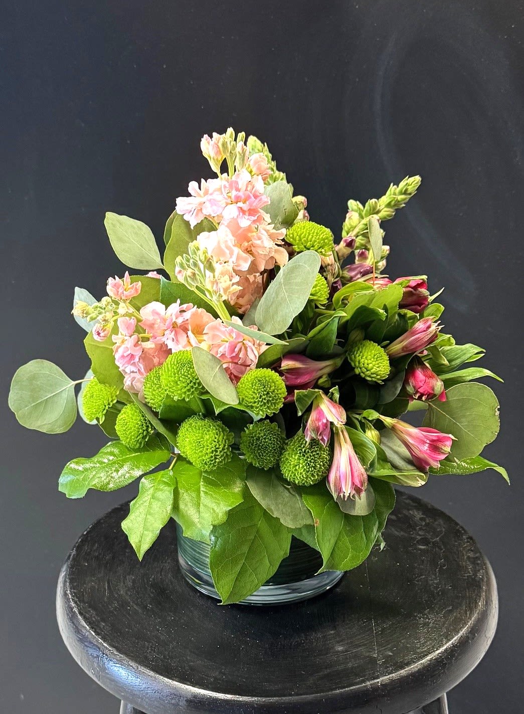 #A103 - Soft Spring Breezes - Soft pink stock takes center stage in this low, compact flower arrangement, its ruffled spires and gentle scent creating a breezy, garden-fresh impression perfect for a spring bouquet or an Easter gift. As a flower arrangement for Mother's Day, a birthday, or a "thinking of you" surprise, it reads as both tender and cheerful-the kind of display that makes the person you're thinking of feel cared for and genuinely seen. Tucked among the stock are bright green button chrysanthemums and pink-and-white alstroemeria, the latter adding delicate stripes and elongated forms that give the design movement and texture.   Densely gathered into a clear glass vase, the blooms form a lush, rounded silhouette with a restorative, close-to-the-garden look. The palette of pink, green, and white works beautifully for occasions from anniversaries and graduations to get well wishes, thank you’s, or simply a just-because pick-me-up. This compact arrangement is easy to place on a bedside table, desk, or coffee table to brighten someone's day-an ideal choice for Mother's Day, Valentine's gestures, weddings, or to say "I'm sorry" with grace. Simple care: refresh water every few days and trim stems as needed to keep the blooms looking their freshest.