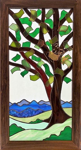 #A104 - Summertime Twin Set (right twin) - Stained Glass Window - A vertical stained-glass panel that reads like a quiet summer morning - this right twin in the Summertime Twin Set is the mirror image of it’s twin, making them perfect to hang in sidelight windows.  This framed stained glass art piece is perfect as a heartfelt Mother's Day gift, a thoughtful housewarming present, or a calming addition for someone you want to encourage. The stylized tree and rolling hills create an immediate sense of peace, so the person you're thinking of will feel seen and comforted every time light plays across the glass. Handcrafted within a wood frame, the panel uses curved brown glass for the trunk and branches and ribbon-like green and yellow-green pieces to suggest fluttering leaves.   The lower scene layers translucent greens for hills, a pale river, and distant blue-purple mountains; the mix of opaque and translucent glass catches daylight differently throughout the day, adding depth and a contemplative glow. Sized to display vertically on a wall or window, it pairs naturally with occasions from anniversaries and birthdays to retirement, sympathy, "I'm sorry," or simple "thinking of you" moments. This piece works year-round-give it for Father's Day, Christmas, a wedding, or "just because" to mark new beginnings-and it especially suits anyone who appreciates quiet, nature-inspired décor. The wood-framed panel arrives ready to hang; no special care is required beyond occasional dusting to keep the glass bright and the scene luminous.