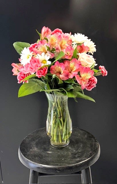 #A106 - Pink &amp; Pretty - Soft pink alstroemeria and ruffled carnations take center stage in this cheerful flower arrangement, a perfect birthday bouquet, Mother's Day gift, or "just because" surprise that reads as warm and thoughtfully feminine. The mix of roses, daisies, alstromeria, and chrysanthemums creates a rounded, slightly airy dome that feels both celebratory and comforting-ideal for anniversaries, get well wishes, new baby visits, or a bright touch for Easter and springtime occasions.   Rendered in pink, white, and touches of sunny yellow, the arrangement is gathered in a clear glass vase so the stems show cleanly and the composition reads fresh from every angle. Pale roses add gentle romance while white daisies and yellow-marked alstroemeria bring contrast and lively detail; the overall size and casual silhouette make it equally at home on a bedside table, desk, or dining centerpiece. Recommend for anyone you want to make feel seen-send it for graduations, housewarmings, thank-you gestures, or to say "I'm sorry" with sincerity. Care is simple: fresh water in the vase and a cool spot away from direct sun keeps the blooms looking their best.