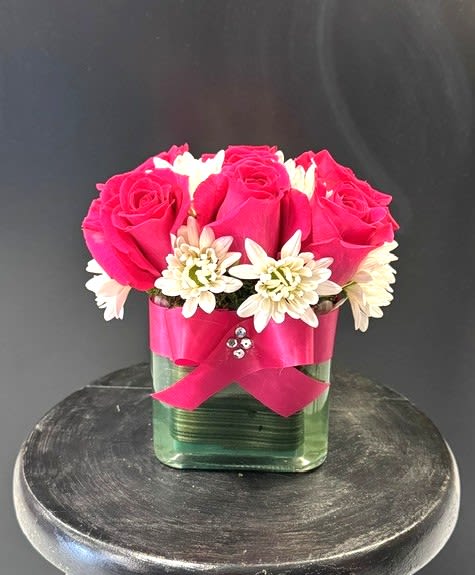 #A007 - Cheerful Vibes - A compact dome of bright pink roses and crisp white chrysanthemums creates an instantly cheerful centerpiece-perfect as a birthday bouquet, Mother's Day gift, or a just-because surprise to brighten someone's day. The arrangement reads feminine and polished, so it also works beautifully for anniversaries, Valentine's Day, or to send thoughtful get-well wishes that feel both lively and comforting.   Flowers are tightly clustered into a low, structured shape that sits neatly in a clear glass cube vase lined with green material for fresh contrast. Daisies mingle with the roses and chrysanthemums for playful texture, while a wide pink satin ribbon folded into an awareness-style loop and tiny rhinestone accents add subtle sparkle and refinement. The low-profile design is ideal for tabletops and desks-an elegant housewarming, graduation, or thank-you gift that suits spring or Easter celebrations but remains seasonally appropriate year-round. 