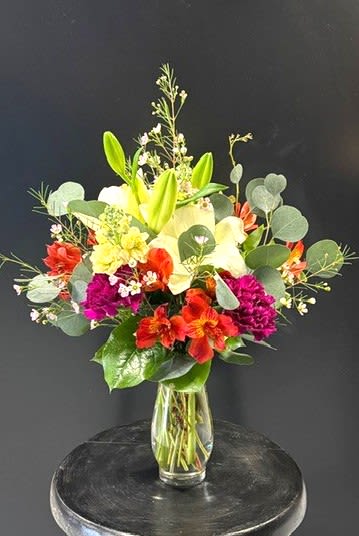 #A019 - Midday Magic - Bright, energetic color and a lively mix of textures make this flower arrangement a midday burst of joy perfect for a birthday, Mother's Day, or just because. The arrangement is a flower arrangement for spring and Easter celebrations as well as year-round gifting-an uplifting choice for Mother's Day, Get Well wishes, housewarming or to say I'm Sorry in a warm, sincere way. The recipient will feel genuinely seen when greeted by that bold contrast of purple, yellow, orange and red.  This design gathers yellow Asiatic lilies, orange-red alstroemeria, rich purple carnations and sprays of white stock and small filler blooms in a clear glass vase. The upright, slightly informal silhouette keeps flowers visible from every angle, making it ideal for a desk, side table, countertop or reception area. The mix of large lily petals and tighter carnation heads creates a pleasing contrast of form and texture that reads cheerful and sophisticated.  Practical for many occasions-anniversary, graduation, thank you, thinking of you, Valentine's Day or retirement-the arrangement arrives ready to display in its clear glass vase. For best longevity, refresh the water every other day and trim stems as needed so the person you're gifting feels that same bright lift for as long as possible.