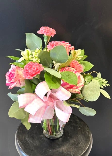 #A074 - New Love - Rounded clusters of bright pink carnations and variegated pink roses give this flower arrangement a cheerful, garden-fresh feel that says "new love" without words. As a birthday bouquet, Mother's Day surprise, or a sweet anniversary or new baby gift, the pink, green, white, and pale yellow palette reads as both romantic and comforting-perfect for Easter or springtime celebrations and just as lovely for thinking-of-you or get-well wishes any time of year. Pale yellow alstroemeria brightens between the rose-and-carnation clusters while abundant rounded green foliage fills the composition for a full, natural silhouette.   The stems are arranged in a clear glass vase and finished with a wide pink plaid ribbon tied into a large bow, lending a feminine, home-ready presentation that makes it easy to place on a bedside table, kitchen island, or office desk. This arrangement is ideal for occasions that call for warmth and gentle cheer-Mother's Day, graduation, housewarming, retirement, or simply to say thank you or I'm sorry. No special care is needed beyond fresh water and keeping the vase out of direct midday sun; it's designed to arrive ready to brighten someone's day.
