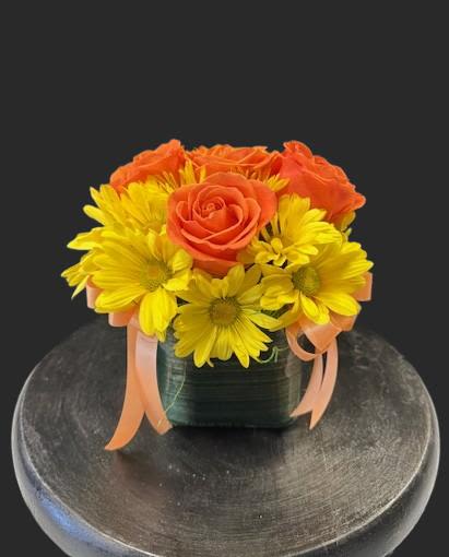 #A133 - Sunshine Cube - Bright, sun-soaked color takes center stage in this cheerful flower arrangement-perfect as a birthday bouquet, Mother's Day surprise, or a bright "get well" pick-me-up. The compact flower arrangement and its low, modern cube vase make it an ideal housewarming or desk-friendly gift for graduations, thank-you moments, or simply to say "thinking of you." Tightly clustered orange roses nestle among vivid yellow daisy-style blooms, creating a bold contrast against fresh green accents.   The stems sit in a leaf-wrapped glass cube vase with soft orange ribbon trails for a playful, slightly whimsical finish. Its clean lines and compact size work well for mantelpieces, office desks, or bedside tables. This sunny arrangement reads as spring and Easter-friendly without feeling seasonal-equally fitting for summer celebrations, anniversaries, new baby welcomes, weddings, or to apologize with warmth. Care is simple: keep water fresh, trim stems if needed, and place out of direct heat to extend the bright, uplifting display.