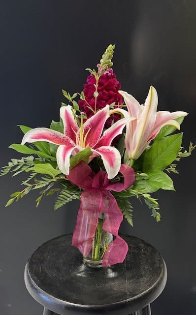 #A015 - Starfighter Vase - A dramatic stargazer-style lily takes center stage in this red, pink and white flower arrangement-an elegant flower arrangement perfect for Mother's Day, anniversaries, birthdays, or simply to say "I'm thinking of you." The wide pink petals edged in white and freckled with darker spots create an immediate visual impact, while deep burgundy snapdragons add vertical drama and contrast that reads romantic and confident at once.  Creamy green foliage frames the blooms and gives the composition depth, all gathered in a clear glass vase finished with a soft pink sheer ribbon bow for a polished presentation. The mix of stargazer lilies and tall snapdragons balances full-bodied texture with graceful height, making it right-sized for tabletops, mantle displays, or a bedside pick-me-up.  Send this arrangement as a bold birthday surprise, a get-well lift, a housewarming or new-baby welcome, or a heartfelt apology-she'll feel genuinely seen the moment it arrives. Arrives arranged and ready to display in its glass vase; trim stems and fresh water will keep the lilies and snapdragons looking their best for several days.