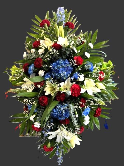 #A047 - Stars &amp; Stripes Standing Spray - Our Stars &amp; Stripes Standing Spray honors our military heroes, first responders, and all those who love this country!  Here at Wild Blooms, we respect everyone's budget so we offer this spray in three sizes: (medium shown in photograph) small for $231.99, medium for $251.99, and large for $271.99.    A bold red, white, and blue standing spray that reads with quiet dignity-perfect for a sympathy or funeral tribute honoring a loved one or a patriotic observance. The elongated teardrop silhouette presents a formal, respectful profile that will make the person you're remembering feel honored and seen. Clusters of bright red carnations are woven with creamy white Asiatic lilies and small white pom-style chrysanthemums for crisp contrast, while rounded blue hydrangea heads and vertical delphinium add depth and a stately blue accent. Lush green foliage frames the composition and lets the gerbera-style shapes and snapdragon lines read clearly from a distance, making this arrangement impactful in a chapel or graveside setting.   Designed as a large standing spray, it's intended to be displayed on an easel where its proportions command attention and convey solemn respect. Care is simple-keep the foam moist until placement and avoid direct heat-so it arrives fresh and ready to make a meaningful statement for the person you're honoring.