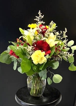 #A083 - Make Someone's Day! - Bright red gerbera daisies and soft yellow carnations take center stage in this flower arrangement, making it a joyful birthday bouquet or a sunny Mother's Day gift that reads as both celebratory and heartfelt. The mix of red, white, yellow and mixed tones-combined with pale asiatic lilies and roses-creates an uplifting color story that immediately tells the person you're thinking of them. Whether you're sending congratulations, a get-well cheer, or a just-because surprise, she'll feel genuinely seen by the warmth and energy of these blooms.  The arrangement is gathered in a clear glass vase for a clean, modern presentation that shows off rounded foliage and delicate white filler blooms framing the brighter flower heads. Texture comes from the layered shapes-bold gerberas, ruffled carnations, open lilies and classic daisies-so it looks full and polished from every angle. Sized to brighten a bedside table, kitchen island, or office desk, it's an easy choice for housewarmings, anniversaries, graduations, or to say "I'm sorry" with sincerity.  Easy-care and long-lasting, the stems arrive arranged and ready to display; change the water every few days and trim stems if you want to extend vase life. Order in time for holidays, retirement celebrations, Memorial Day remembrances, or a simple thank-you-this versatile bouquet brings warmth to spring, summer and autumn occasions alike.