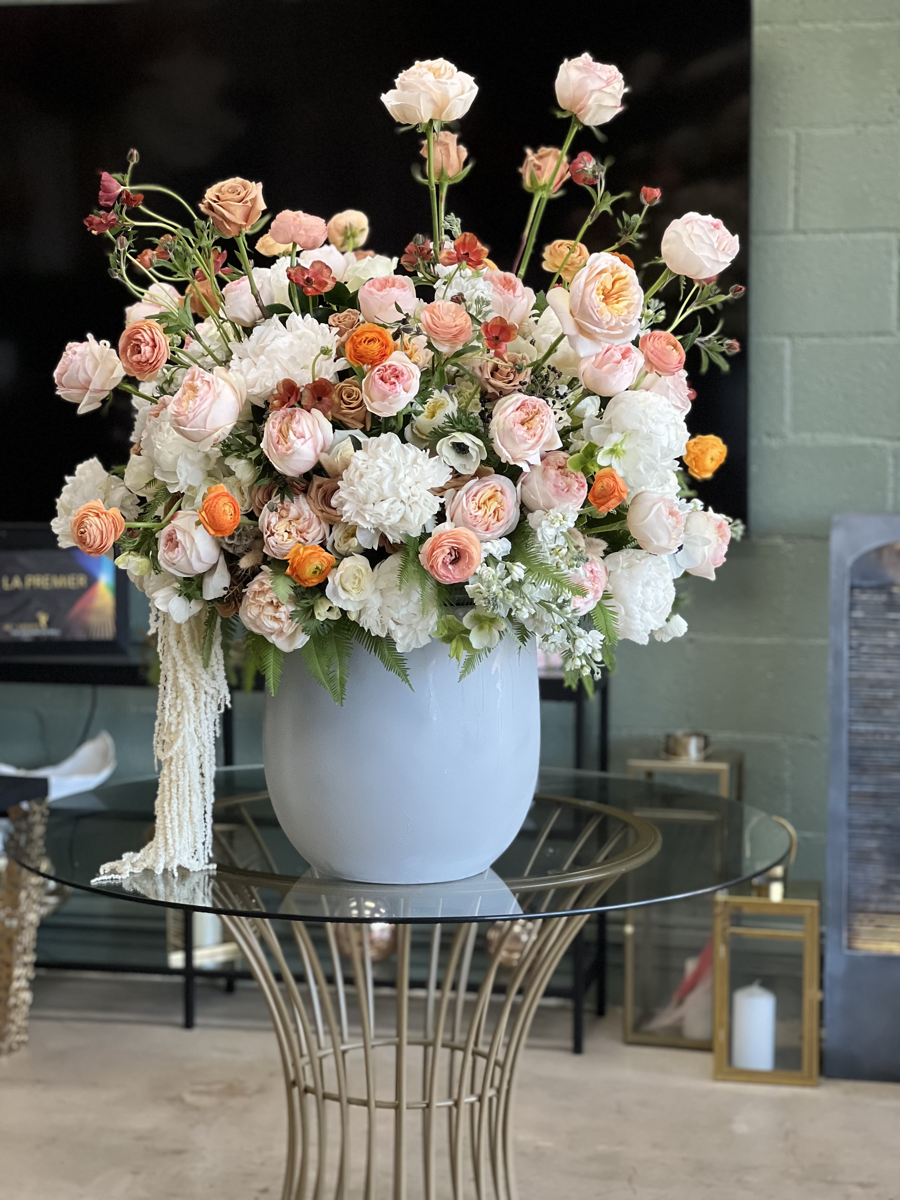Zuley Pastel Garden Rose and Peony Arrangement - Lush pastel blooms in a smooth white ceramic vase make an unforgettable birthday bouquet or anniversary centerpiece - Zuley is a flower arrangement that reads like a soft garden in bloom. The layered mix of pink, white, peach, and soft orange gives the person you're celebrating a feeling of warmth and tenderness, perfect for saying "thinking of you," "congratulations," or "happy birthday" and equally lovely for a romantic anniversary table or a thoughtful get well gift.  This large, abundant centerpiece pairs garden roses, peonies, anemone, ranunculus and roses with fuller white blooms for contrast; additional daisies and hydrangea tones appear in photos for extra texture. Long stems and scattered blossoms rise above a dense, rounded base to create height and movement while the smooth white ceramic vase keeps the look elegant and wearable for any room. Sized to fill a table or entryway, Zuley arrives ready to display on a glass table or mantel - it's the kind of arrangement that makes the recipient feel genuinely seen, whether you're marking a milestone, brightening a new baby celebration, saying sorry, or surprising someone just because in Los Angeles or beyond.