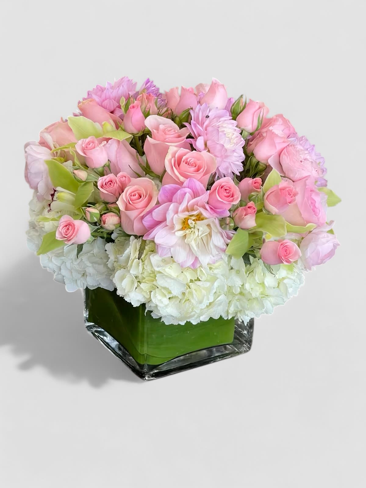 Mandala - A simple pavé of pink Roses, Dahlias, and green Cymbidium Orchids on a bed of white Hydrangea, in a leaf-lined glass vase