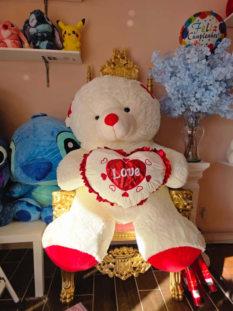 Large teddy bear holding a heart pillow beside a vase of blue flowers