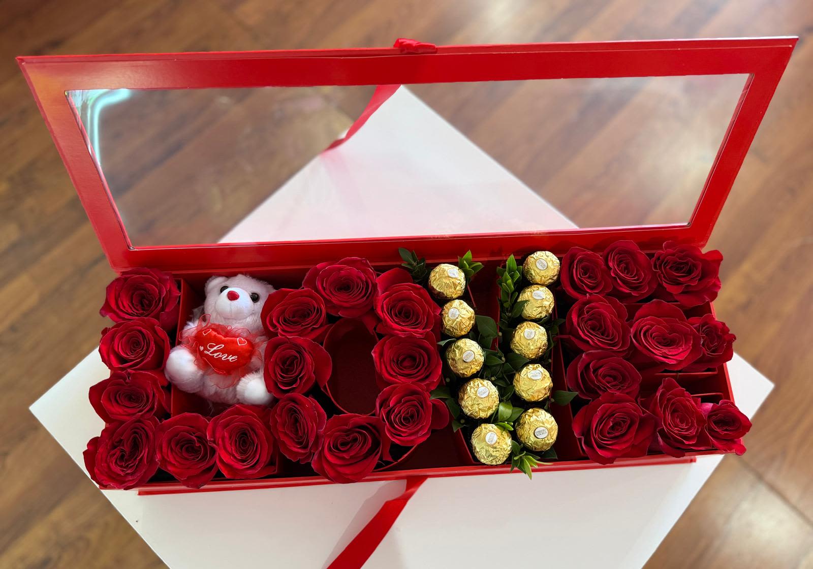 Red roses arranged in a red gift box with a small teddy bear and chocolates