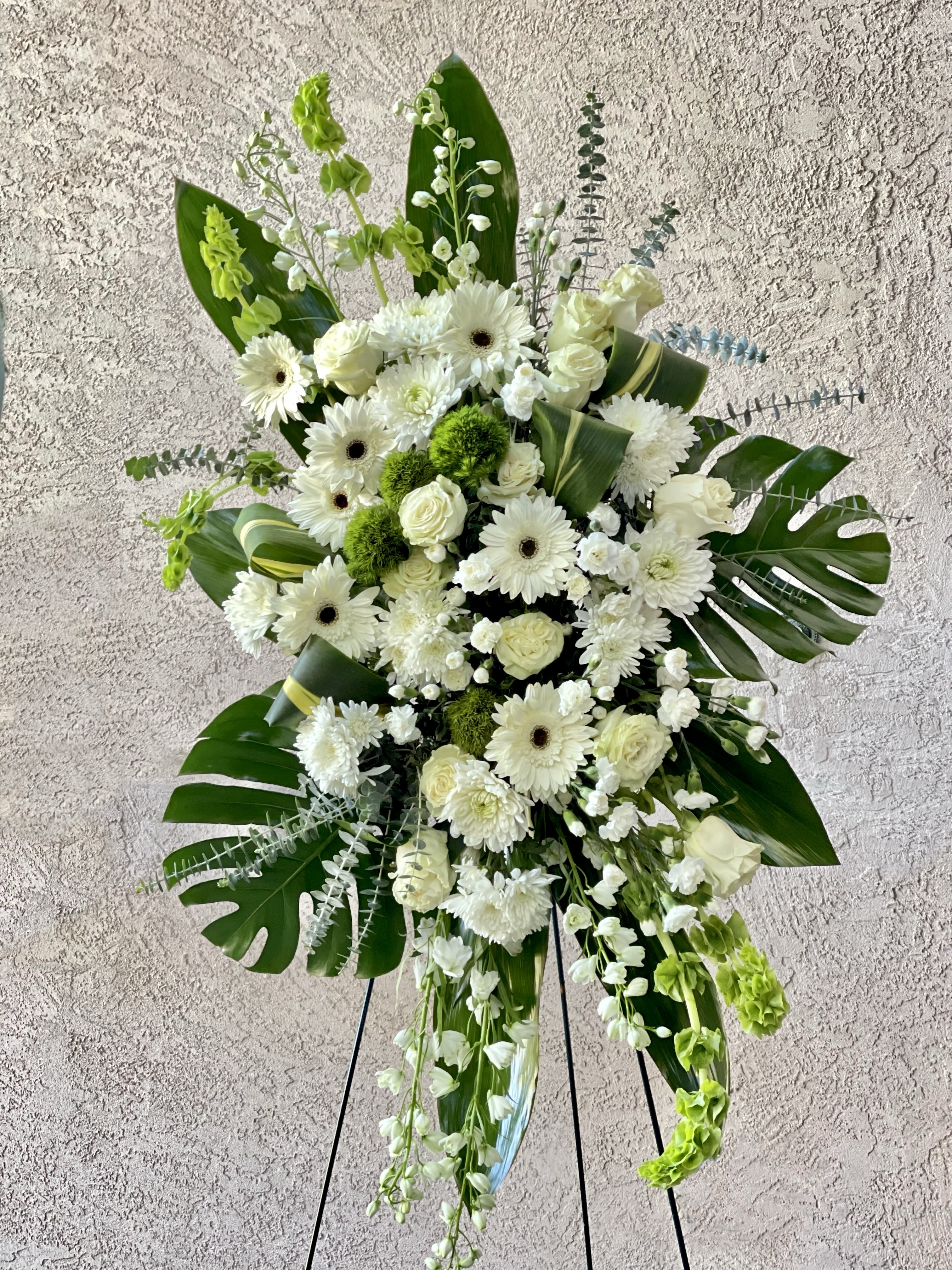 Tall standing spray of white roses, gerbera daisies, and mums with large tropical leaves on an easel