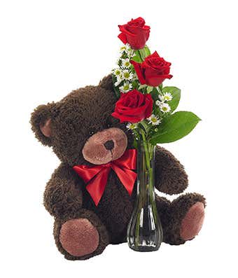 Brown teddy bear with red bow beside a clear vase of red roses and small white daisies.
