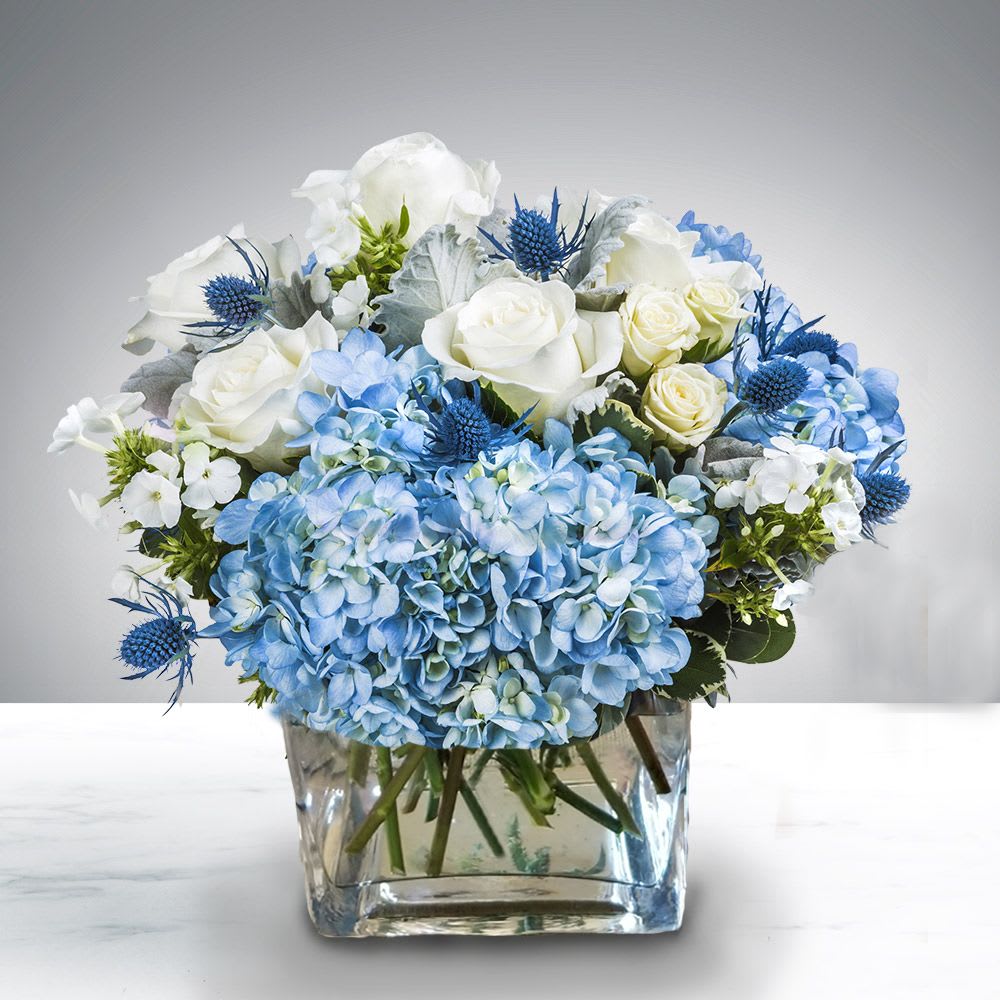 Low glass vase of blue hydrangeas, white roses, and small accent blooms in blue and white.