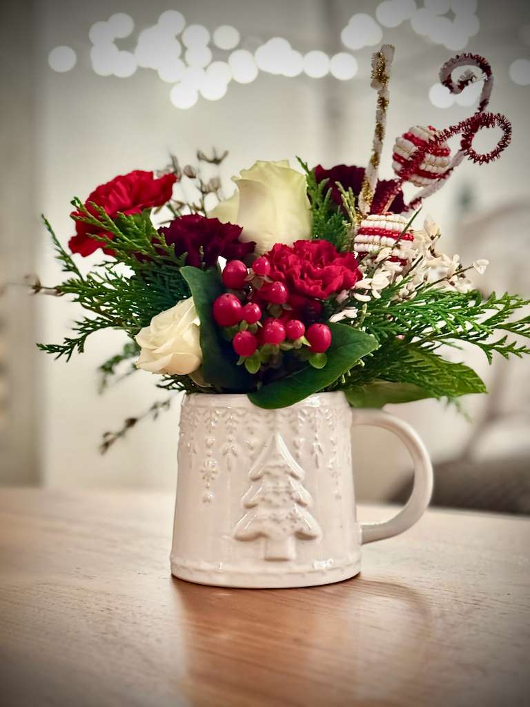 Christmas Mug Floral Arrangement – Limited Edition - Our Christmas Mug Floral Arrangement is a charming seasonal design created in a festive holiday mug and styled with classic Christmas tones, seasonal greenery, and delicate details.  Perfect for small gifts, teacher appreciation, hostess gifts, or adding a cozy holiday touch to any space.  Each arrangement is handcrafted with care and offered in limited quantities to ensure quality and uniqueness.  Seasonal flowers and mug styles may vary based on availability while maintaining the overall holiday style and color palette