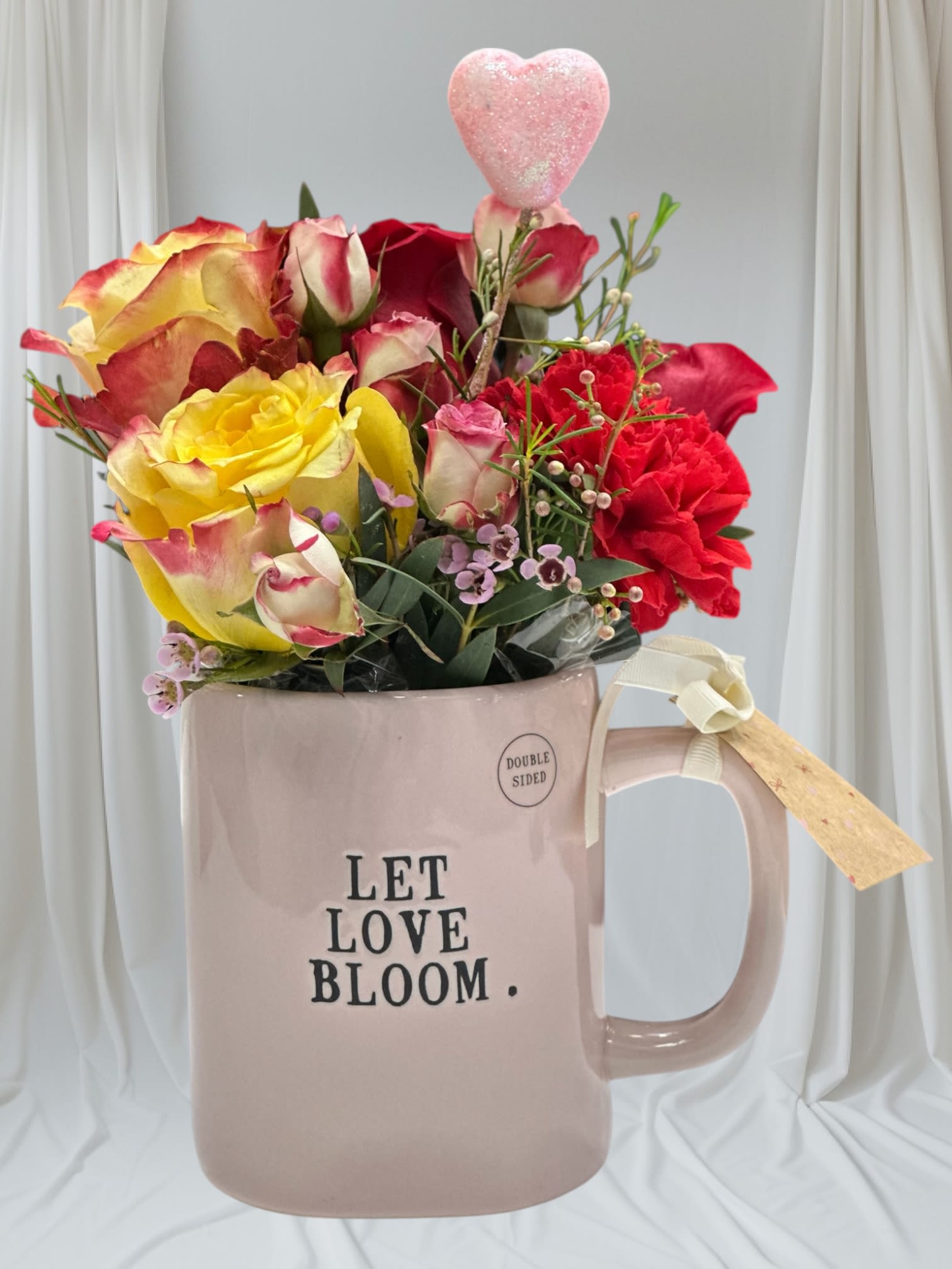 Let Love Bloom Mug Arrangement  - A romantic Valentine’s Day floral arrangement designed in a keepsake mug that reads “Let Love Bloom.”  Handcrafted with fresh seasonal flowers in shades of red, pink, and soft yellow, accented with delicate greenery and a sweet heart detail. Perfect as a thoughtful Valentine’s gift or a charming gesture of love.  Each arrangement is made by hand with care, making every piece unique.