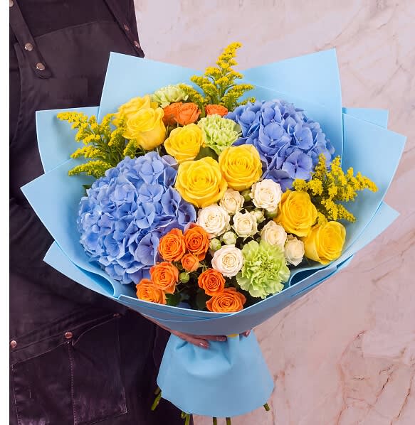 Bouquet of blue hydrangeas, yellow and orange roses, and green carnations in blue wrap
