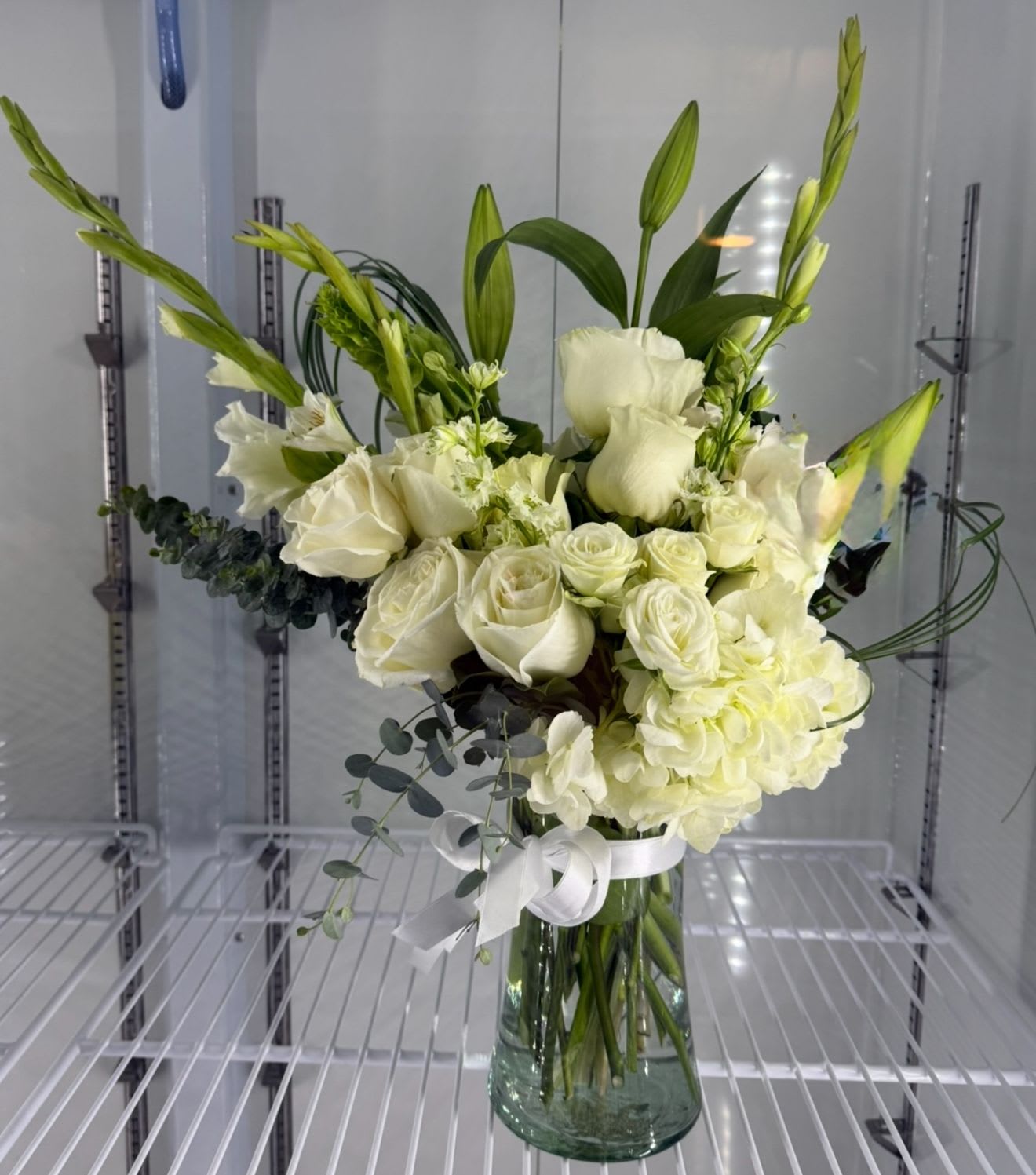 Tall arrangement of white roses, lilies, and hydrangea with green buds in a clear glass vase.