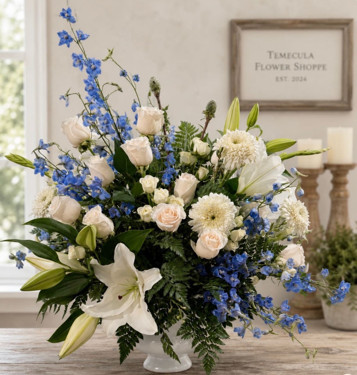 Blue delphinium, white lilies, and cream roses in a white pedestal vase