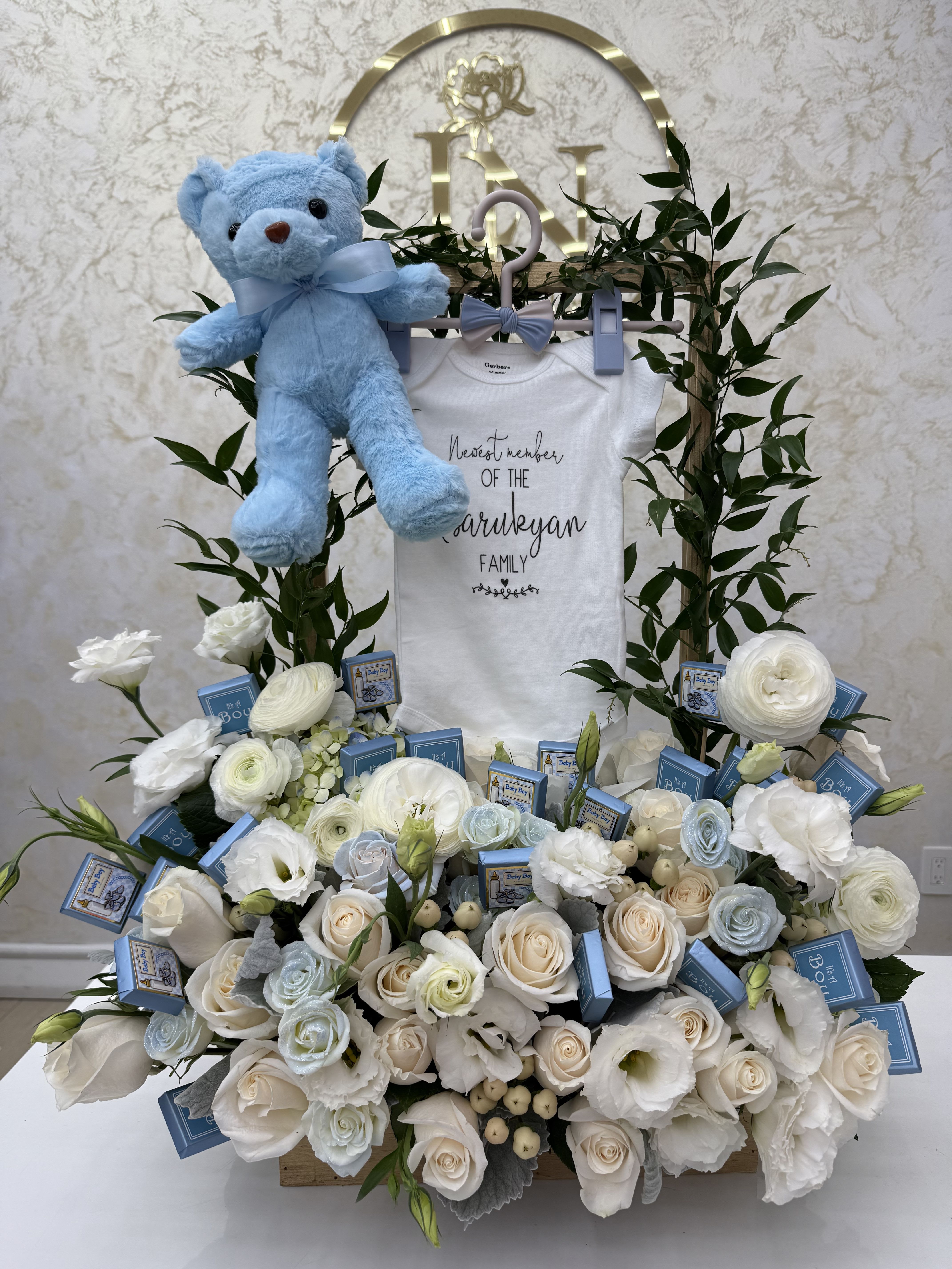 White floral baby gift arrangement with a blue teddy bear and baby onesie