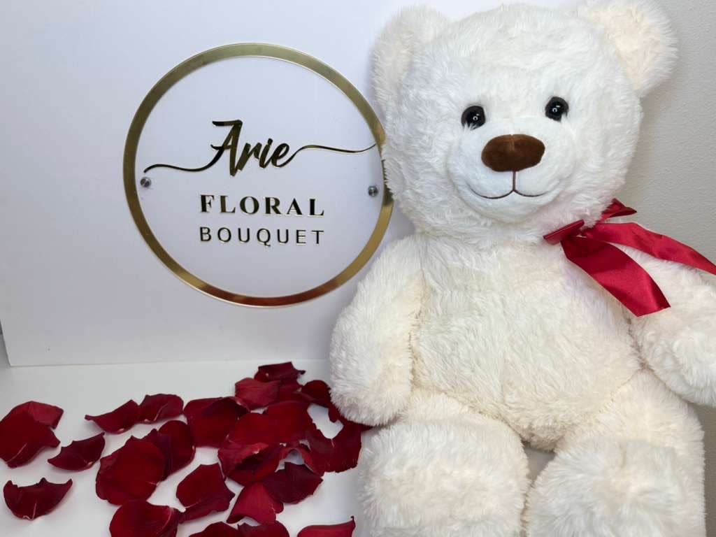 White teddy bear with a red ribbon beside rose petals and a floral bouquet sign