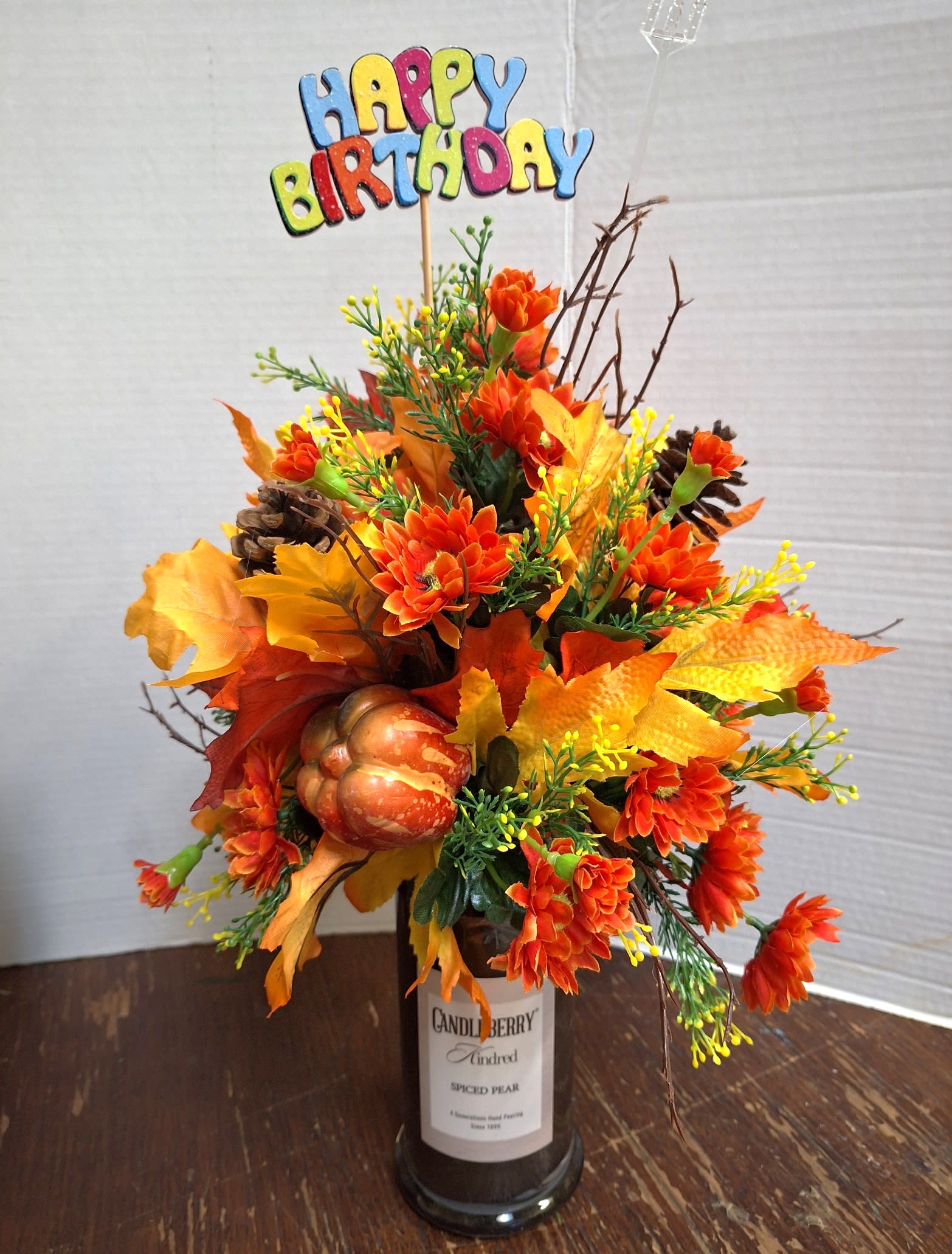 Floral Scents Candle - Bright orange silk chrysanthemum-style blooms take center stage in this cheerful birthday arrangement, styled to read like a decorative candle. The flower arrangement and occasion - birthday bouquet for fall celebrations - are clear from the colorful "Happy Birthday" pick that tops the display, so the person you're thinking of will feel celebrated the moment they see it.  Crafted in a labeled glass candle jar, the compact arrangement mixes orange and yellow faux leaves with textured pinecones, a small artificial pumpkin, and thin decorative branches for height and movement. The silk chrysanthemums are densely clustered for full visual impact; the glass jar gives it a clean, gift-ready finish that fits on tabletops, mantels, or kitchen counters.  Perfect for fall birthdays or anyone who loves autumnal style, this piece is low-maintenance and long-lasting since the flowers are silk and the accents are all decorative. Sized for easy placement, it arrives ready to display - no water or care needed - making it an effortless, festive way to make someone feel truly seen on their special day.