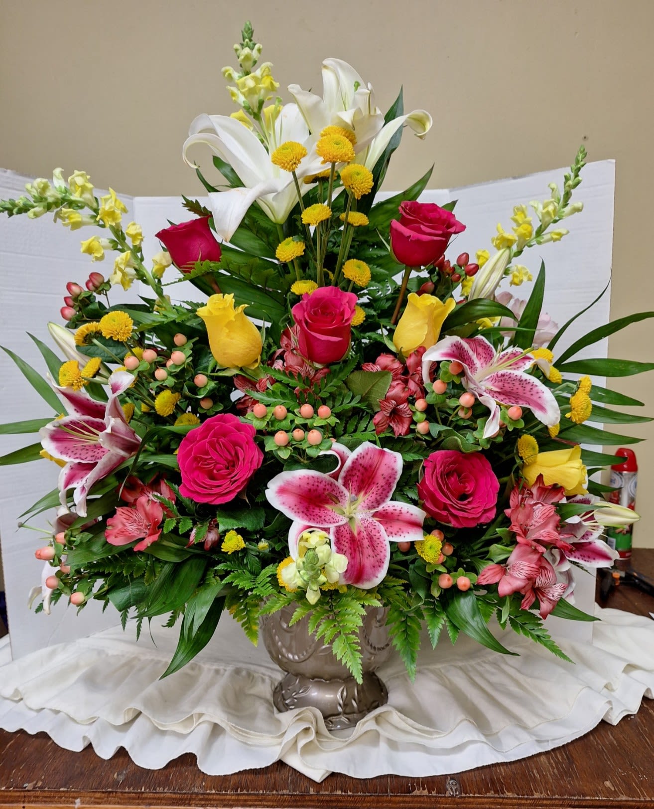 Colorful Cascade - A tall, fan-shaped sympathy and funeral arrangement that reads as both comforting and uplifting-the Colorful Cascade brings warmth with pink, white, yellow and mixed-color blooms arranged in an ornate plastic urn container. This funeral flower arrangement and sympathy piece uses its bright palette to celebrate a life, while also offering a gentle, hopeful presence that works for memorial services or to send condolences.  The design features abundant pink roses and yellow roses paired with speckled pink stargazer lilies and Asiatic lilies for a layered, fragrant focal. Pink alstroemeria and clusters of small yellow button chrysanthemums add dense color and texture, while yellow and white snapdragons lend vertical rhythm and a graceful, fan-like silhouette. Berry-like accents tuck between blooms for depth; the flowers are set in a classic ceramic urn that gives the piece a traditional, evergreen feel.  Ideal for honoring a loved one at a funeral service or sending heartfelt sympathy that feels celebratory rather than somber, this arrangement also suits birthdays, get-well wishes, or congratulations when a vibrant, supportive message is called for. Typical sizing fills a ceremony space with a prominent presence; please contact us for delivery timing or special requests to ensure the arrangement arrives fresh and properly staged.