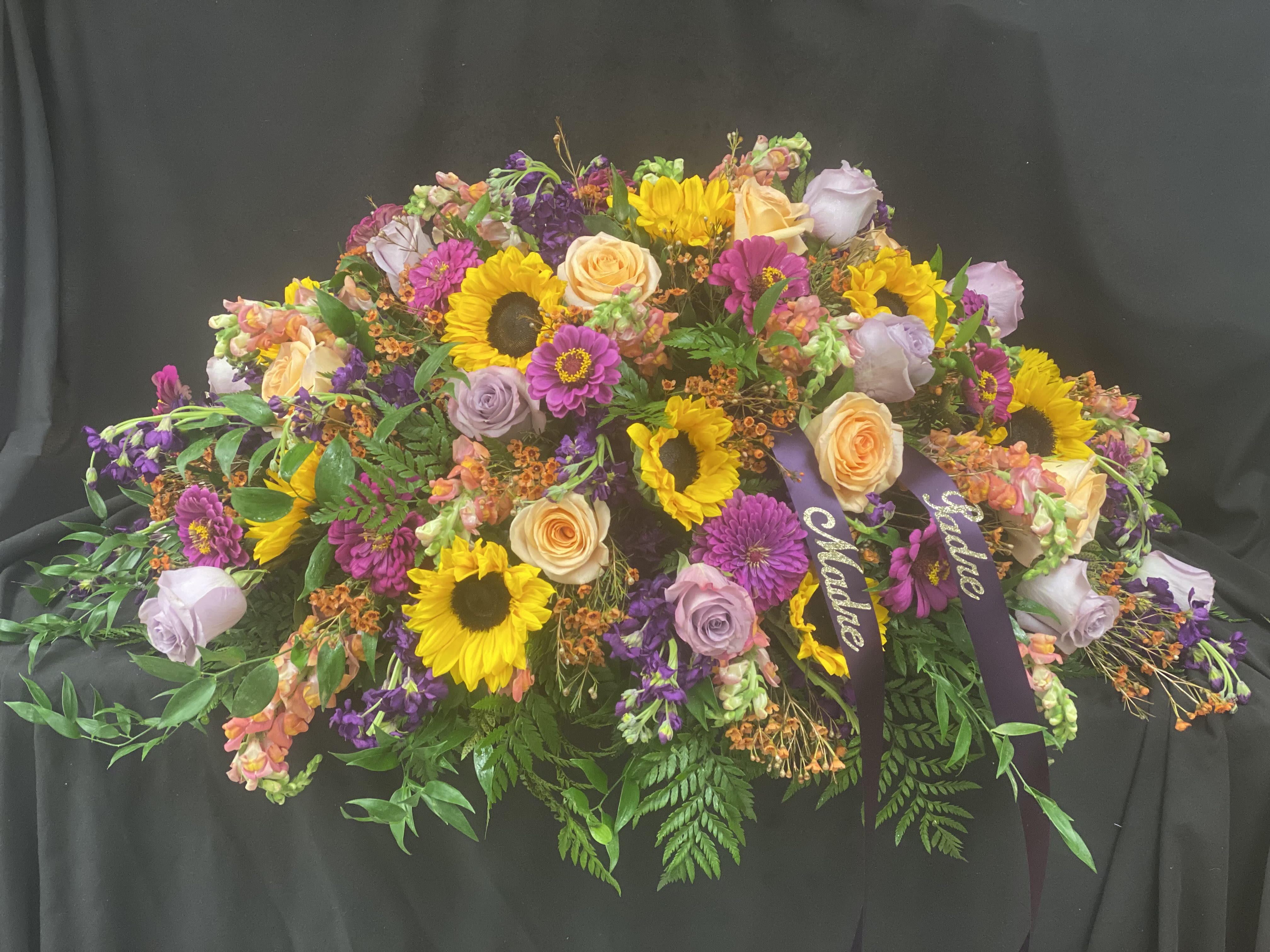 Casket spray with sunflowers, roses, and purple blooms on a dark drape