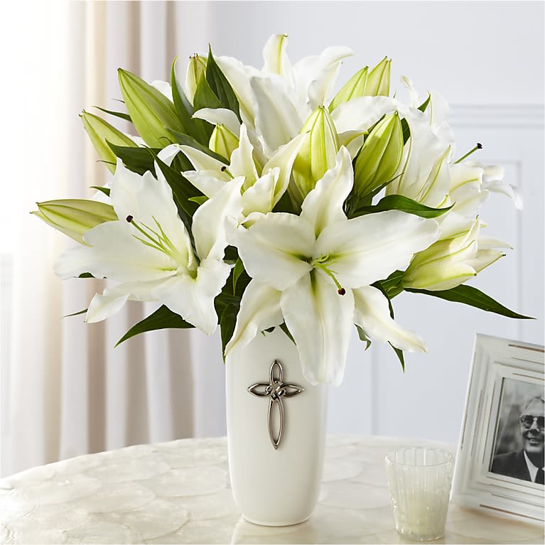 White lilies arranged in a tall white vase with a cross emblem