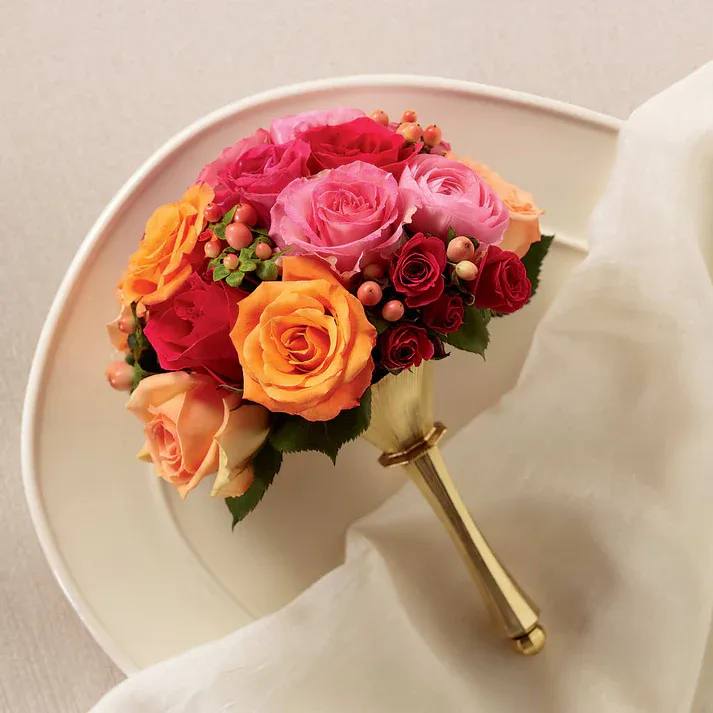 Bright Promise Bouquet - Our Bright Promise Bouquet is a combination of captivating color and stunning bridal elegance to create the perfect wedding day look. Orange, peach, pink, and fuchsia roses are arranged amongst fuchsia spray roses, pink ranunculus and peach hypericum berries, all brought together in a gold metallic bouquet holder to create a bouquet of charm and grace.  Standard bouquet is approximately 11" H x 8" W