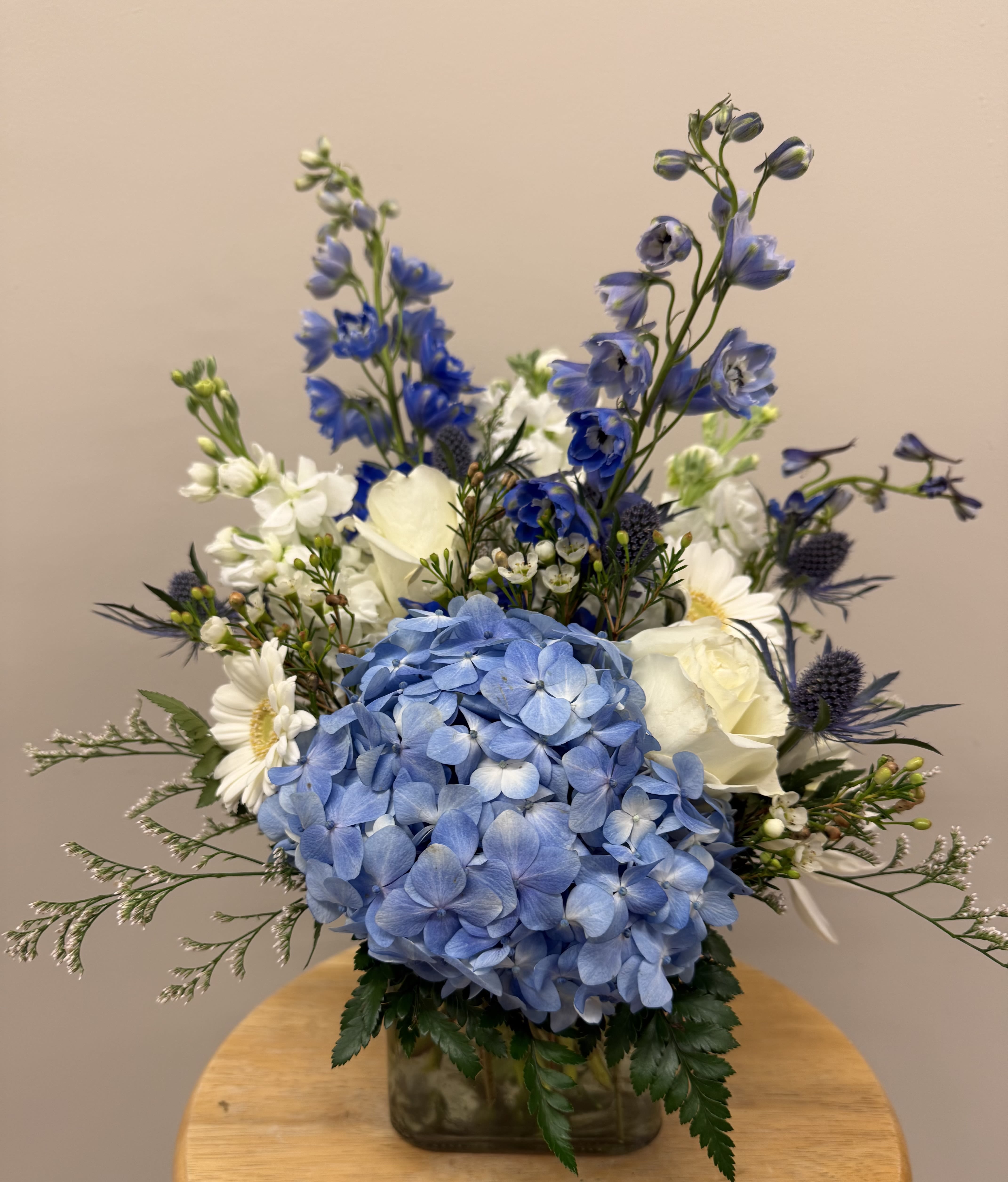 Blue Haze - A lush blue hydrangea takes center stage in this Blue Haze flower arrangement, its cloudlike bloom surrounded by crisp white roses and cheerful white daisy-style flowers for a serene, garden-fresh look. Tall blue delphinium spikes add graceful height while small purple thistle-like accents bring a touch of texture-making this arrangement an ideal birthday bouquet, thinking-of-you gift, or springtime surprise for someone who appreciates cool, calming colors.  Arranged in a clear square glass vase, the composition balances rounded hydrangea and rose forms with airy delphinium spires to create layered depth that reads both fresh and slightly formal. Colors are strictly blue, white, and purple, and the mix of hydrangeas, roses, daisies, delphinium, and filler blooms gives a natural, sophisticated silhouette that fits well on a mantel, desk, or dining table.  Perfect for expressing thanks, sending sympathy, or simply letting someone know you're thinking of them, this arrangement arrives ready to display in its glass vase. For best results, refresh the water every other day and trim stems slightly if needed to keep the blooms looking their calm, collected best.