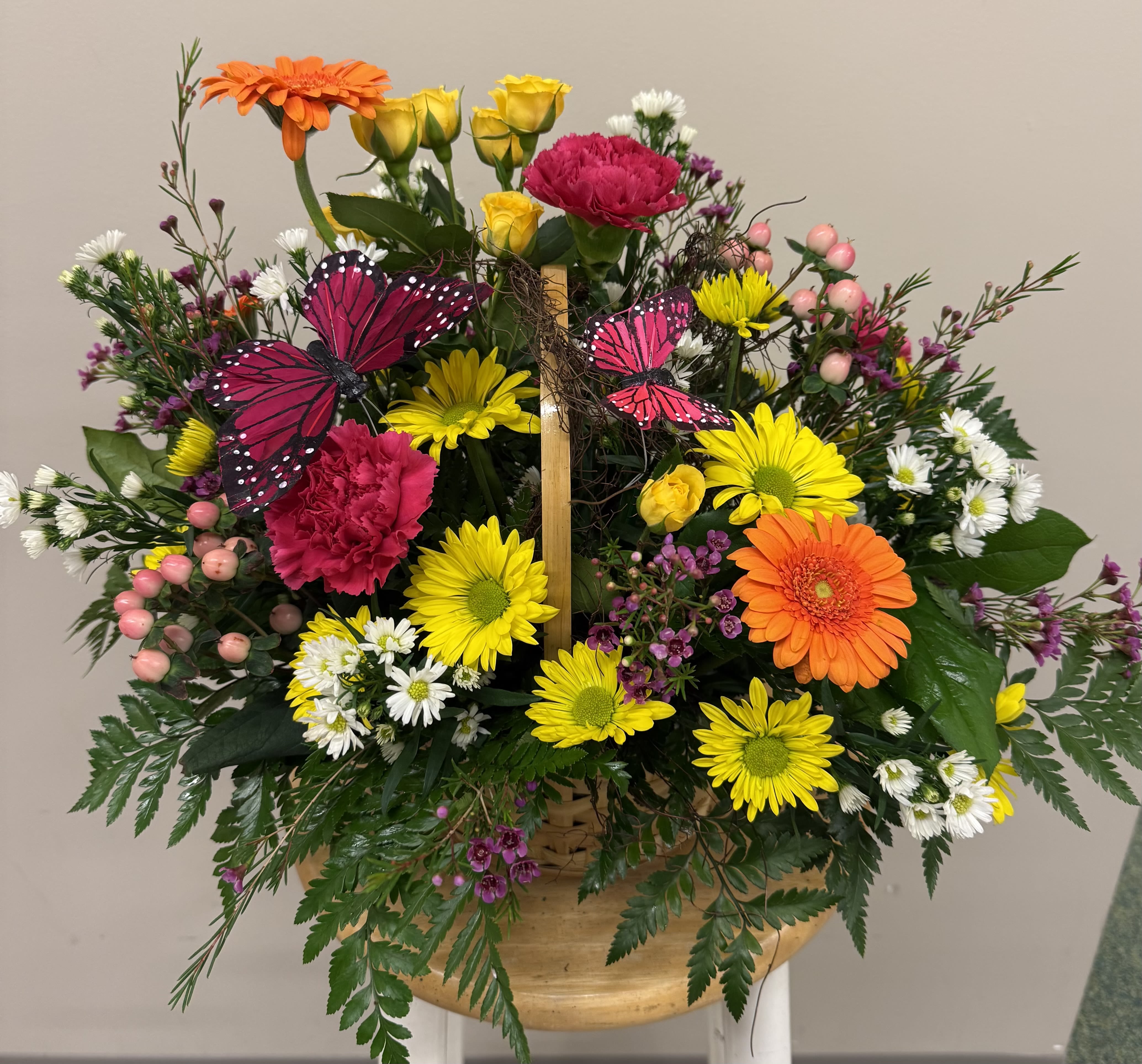 Butterfly Fields - A lively burst of spring in a woven basket-this flower arrangement is a joyful mixed bouquet perfect for a birthday bouquet, a "thinking of you" gift, or a cheerful get-well surprise. The design features bright gerbera daisies and sunny yellow roses up front, with pink carnations and white daisy-like blooms layered throughout to create an energetic, celebratory look. Decorative pink-and-black butterfly picks lend a whimsical, garden-party feel that will make the recipient smile.  Arranged in a roomy woven basket with a wooden handle, the composition is dense and full, framed by abundant greenery for natural texture and contrast. You'll find gerbera daisies, roses, carnations, chrysanthemums and small daisies combined for a variety of shapes and petal textures-bold centers, ruffled petals, and delicate sprays-that read beautifully from every angle. The color palette of pink, yellow, orange and white keeps the bouquet bright and versatile for spring, Mother's Day, or anytime you want to lift someone's spirits.  This is the kind of arrangement that makes someone feel genuinely noticed-playful, warm, and celebratory without being over-the-top. It arrives ready to display in the basket; keep flowers in fresh water, away from direct heat, and recut stems if you transfer them to a vase to extend bloom life. Order now for same-week delivery to make a sunny impression on the person you're celebrating.