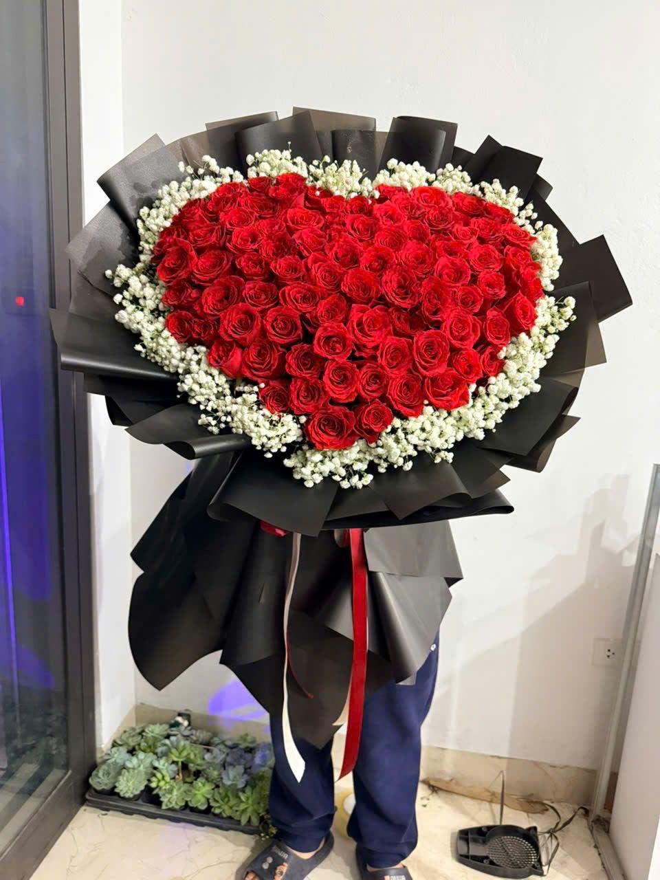 ramo bouquet de corazon By Special Moment´S - A bold heart-shaped arrangement of densely packed red roses edged with delicate white filler-this flower arrangement is a romantic bouquet perfect for an anniversary or Valentine's Day. The dramatic red-and-black palette and sculpted heart form make the recipient feel deeply cherished and seen, the kind of gift that reads as intentional and unforgettable.  Wrapped in striking black paper and finished with a trailing red ribbon, the bouquet contrasts vivid blooms against dark, textured wrapping for high visual impact. The centerpiece is all red roses with clusters of small white accent flowers along the heart's edge; the sturdy wrap keeps the shape crisp and ready for presentation. Ideal for expressing love or deep admiration, this arrangement arrives prepared to give-a bold, show-stopping way to say "te quiero" on a special occasion.
