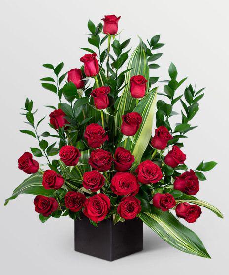 arreglo floral By Special Moment´S - A dense cluster of velvety red roses rises from a sleek black square vase to create an upright, structured statement perfect for a romantic birthday bouquet, anniversary roses, or a Valentine's Day gift. The concentrated mass of blooms with a few stems lifting above the main group gives the arrangement a layered, garden-fresh silhouette that reads both classic and intimate-exactly the kind of floral gesture that makes the person you're thinking of feel noticed and cherished.  Glossy, tall greenery and long variegated leaves add vertical drama and polished contrast to the rich red petals and deep green foliage, while the black square vase frames the colors for a modern, formal finish. The composition is full and compact with roses focused at low and mid levels for a lush visual impact; the overall look is ideal for a dining table centerpiece, office desk, or bedside display. This arrangement arrives ready to present-no extra trimming required-and suits everyday gifting as well as milestone moments when you want your recipient to feel truly celebrated.