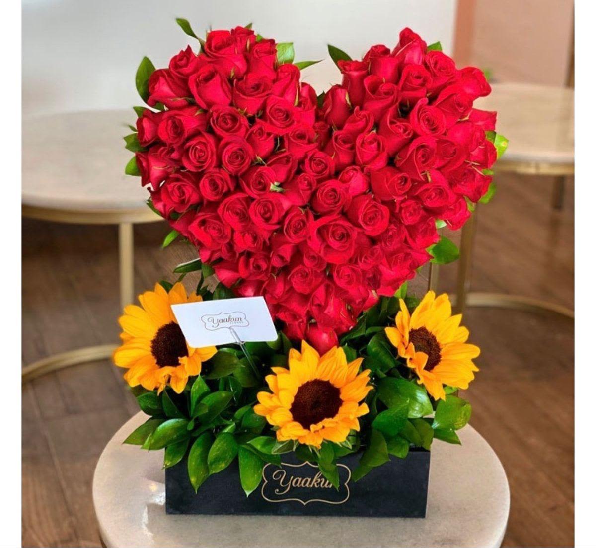arreglo de corazon con R y G By Special Moment´S - A bold heart of vivid red roses rises above a sunny base of bright yellow sunflowers in this flower arrangement-an eye-catching heart-shaped bouquet perfect for an anniversary, Valentine's Day, birthday, or a romantic "just because" gift. The contrast of passionate red and cheerful yellow creates an immediate emotional impact: she'll feel adored and celebrated the moment she sees it.  The roses form a dense, sculpted heart set into a sleek black rectangular box, with glossy green foliage filling and framing the lower portion for a crisp, modern look. Sunflowers fan out beneath the heart, adding warmth and texture while a small card and branded label finish the presentation. Colors are rich-red, yellow, green and the signature black of the box-making this arrangement both decorative and gift-ready.  This design is ideal for someone you want to make feel truly seen-romantic partners, close friends marking a milestone, or anyone who loves bold color. Arrives assembled in the black box with a card for your message; keep flowers in a cool spot out of direct sun and refresh water as needed to extend their life.