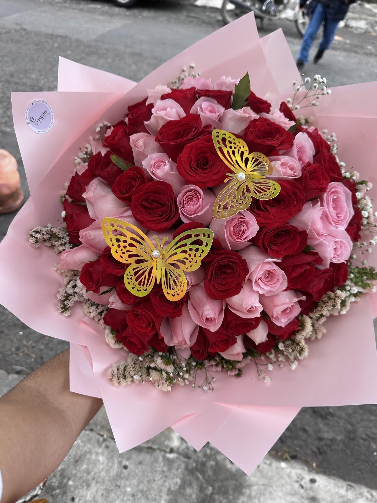 ramo bouchon de rosa y maripoza By Special Moment´S - A lush cluster of red and blush-pink roses creates an immediately romantic centerpiece-perfect as an anniversary bouquet, Valentine's Day gift, or a birthday surprise that says "you're cherished." The rounded, full shape feels abundant and feminine, with white filler blooms softening the edges and two gold butterfly decorations adding a playful, celebratory touch she'll notice.  Wrapped in pale pink paper that frames the blooms like a soft ribbon, this arrangement blends red, pink, white and a hint of yellow in its palette (subtle yellow accents lift the mix). Dense rose heads give a plush, tactile feel and a classic rose scent; the compact, rounded design makes it easy to hold and display on a table or bedside. For the person you want to make feel truly seen, this bouquet delivers warmth and romance in a gift-ready presentation-order with standard sizing and expect a full, hand-tied arrangement as pictured.
