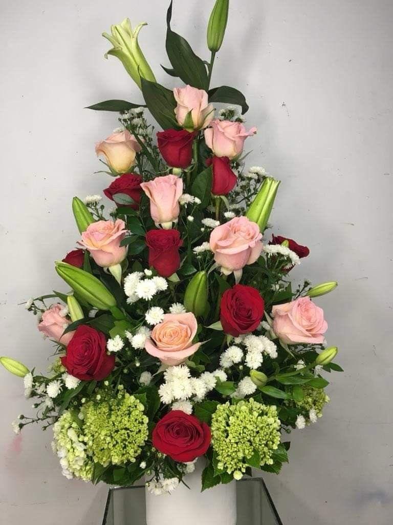 arreglo de rosas y lilys By Special Moment´S - Tall, tiered red and blush roses mingle with unopened lily buds to create a romantic birthday bouquet or anniversary flowers that feels both classic and thoughtfully composed. This flower arrangement for celebrations or heartfelt moments is arranged in a formal, structured silhouette so the person you're thinking of will feel genuinely seen and celebrated the moment it arrives.  The design combines red and pink roses, white lilies (several buds), small white filler blooms, and rounded green hydrangea clusters for texture and volume near the base. Built in a clean white vase, the piece stands upright with layered height for a striking centerpiece effect; the mix of open roses and closed lily buds keeps the look fresh as the lilies gradually unfurl.  Ideal for anniversaries, birthdays, or as a romantic surprise, this is the kind of arrangement that reads as refined and intentional - perfect for someone who appreciates classic floral lines. Care is simple: keep the vase topped with fresh water and place in a cool spot away from direct heat to extend the life of the roses and encourage the lilies to open over the next few days.