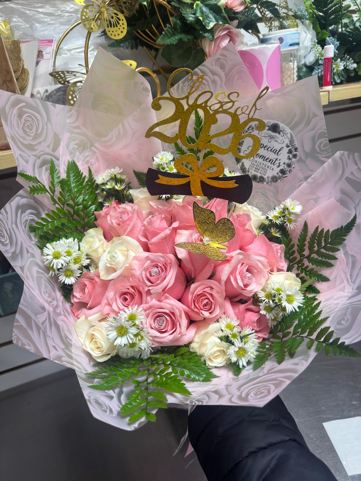 ramo bicho By Special Moment´S - A joyful cluster of pink and white roses makes a memorable graduation bouquet-perfect as a graduation bouquet or celebratory gift for the Class of 2026. The ramo bicho By Special Moment´S is designed to make the person you're honoring feel noticed: soft pink roses at the center, a handful of whisper-white roses, and delicate daisy-like white fillers combine for a feminine, uplifting look that reads "congratulations" at a glance.  Wrapped in translucent pink and gray patterned paper, the stems are presented with a layered, polished finish that elevates the blooms. A gold glitter topper that reads "Class of 2026" and a charming butterfly pick add sparkle and playful detail, while touches of green foliage give contrast and texture. The bouquet reads cheerful and refined-great for a daughter, friend, graduate, or anyone celebrating a milestone.  Give this to the graduate who loves romantic pinks and pretty details; it's the kind of bouquet that makes them feel celebrated and seen. Packaged ready to present, keep the roses in fresh water and out of direct sun to prolong their vase life.