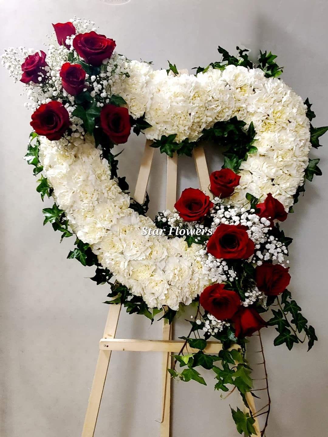 corazon clavel y rosas By Special Moment´S - A large heart of pure white blooms edged with deep red roses creates a powerful, reverent statement-the perfect sympathy arrangement or memorial tribute to honor a loved one. This heart-shaped funeral spray on a wooden easel reads as both romantic and solemn, making it suitable for a celebration of life, graveside service, or memorial ceremony.  Dense white carnations and other white flowers form the body of the heart, while clusters of red roses punctuate the outer edge; small white filler flowers add delicate texture and dark green ivy-like foliage outlines the shape and trails gracefully down. Mounted on a sturdy wooden easel for upright display, the piece is designed for visibility and lasting presence at a service.  Choose this arrangement when you want the person you're remembering to feel deeply honored-its classic white-and-red palette conveys love, respect, and remembrance. Careful delivery and placement on the easel ensure it presents beautifully at viewing or service; please let us know service details for timely delivery.