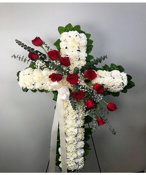 cruz clavel y rosas By Special Moment´S - A dignified white cross formed from dense carnations, accented with deep red roses, creates a reverent sympathy arrangement perfect for funeral and memorial services. This standing flower arrangement for remembrance combines the visual solemnity of white blooms with the heartfelt contrast of red roses, the kind of tribute that helps the person you're honoring feel truly remembered.  Designed on a standing easel, the cross is outlined with dark green foliage and finished with a white ribbon draping from the lower center for a classic, devotional look. The carnations provide full, textured coverage while the red roses are placed across the front and diagonally through the design to draw the eye and add emotional warmth. Sized as a large standing piece, it's intended for use at services, viewings, or graveside tributes; no special care is required beyond keeping blooms cool and shaded until placement.