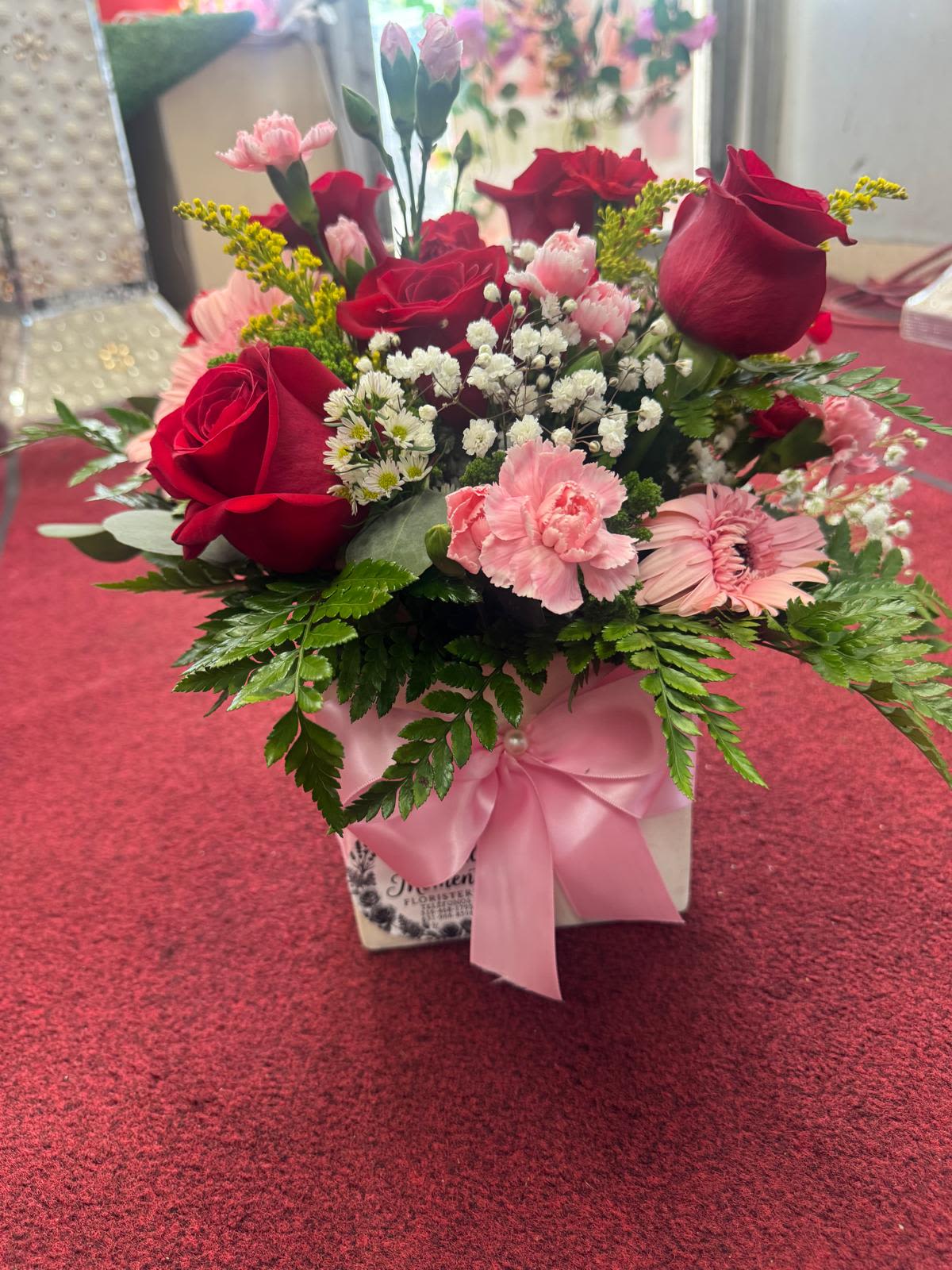 caja de rosas y clavel By Special Moment´S - Deep red roses and soft pink carnations combine with pale pink gerbera daisies in a compact, rounded arrangement-perfect as a romantic bouquet or a cheerful birthday flower arrangement. Presented in a white square gift box tied with a wide pink satin ribbon, this flower arrangement for love and romance or a thank-you moment makes the person you're thinking of feel adored and celebrated at first sight.  The mix includes rich red roses, blush pink carnations, pale pink daisies and small white filler blooms set against fresh greenery for contrast. The low, boxed design keeps blooms closely gathered for maximum visual impact and a polished presentation that travels well as a gift.  Ideal for anniversaries, Valentine's Day, birthdays, or simply to say "thinking of you," this boxed arrangement arrives ready to place on a table or desk-no vase required. For best enjoyment, keep in a cool spot away from direct sunlight and replenish water as needed to extend the life of the flowers.