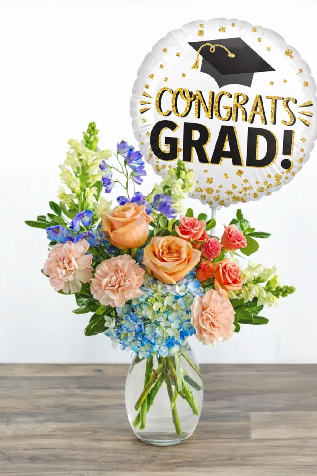 Congrats Grad Floral Arrangement - Celebrate their big achievement with a bright and elegant Graduation Floral Arrangement designed to make their moment unforgettable. This stunning design features a beautiful mix of soft peach roses, delicate hydrangeas, cheerful snapdragons, and seasonal blooms in fresh, uplifting tones.  Arranged in a clear glass vase for a clean and timeless look, this piece brings together color, texture, and height—making it perfect for gifting or displaying at any graduation celebration. Topped with a festive “Congrats Grad!” balloon, (Balloon Color May Vary depending on Availability) it’s a complete gift that delivers both beauty and joy.   Perfect for:  Graduation day surprises Party centerpieces Gift deliveries Celebrating any graduate  Thoughtfully designed to capture the excitement of new beginnings, this arrangement is a meaningful way to say “you did it!” 