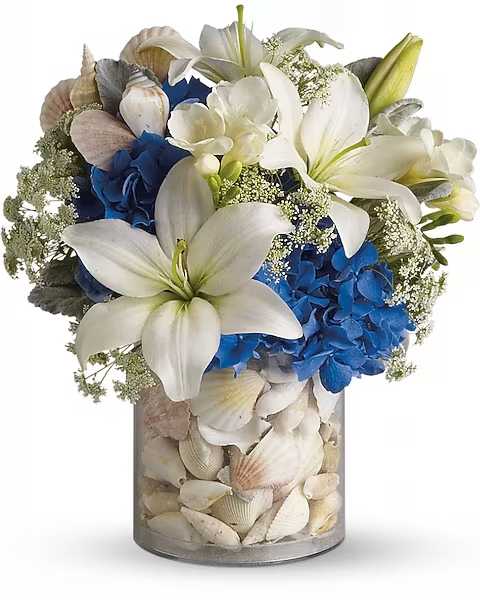 White lilies and blue hydrangeas in a shell-filled glass vase