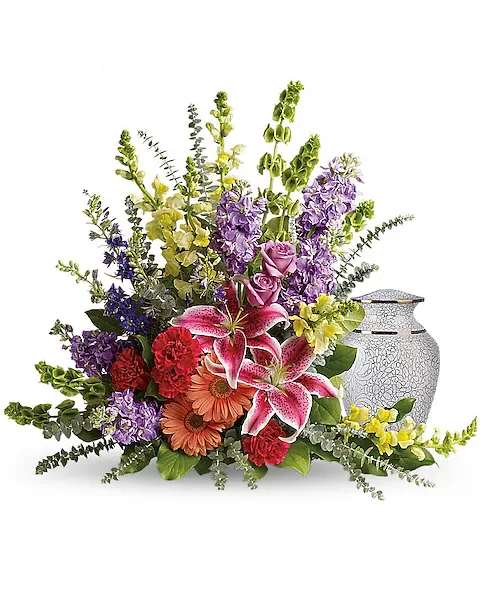 Colorful floral arrangement with lilies, roses, and gerbera daisies beside a white urn