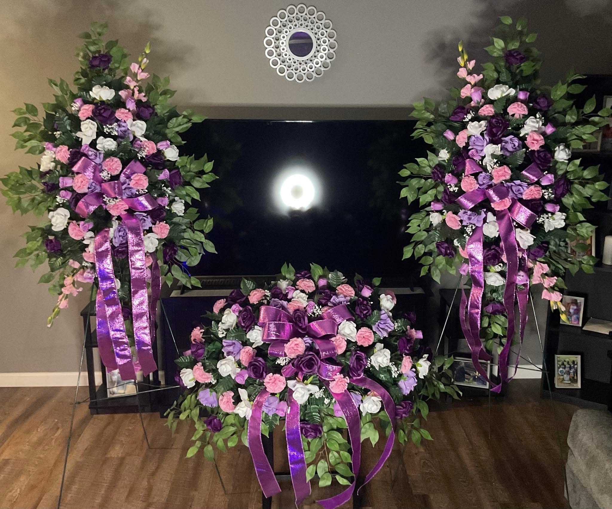 Three purple, pink, and white floral sprays with ribbon accents arranged around a TV.