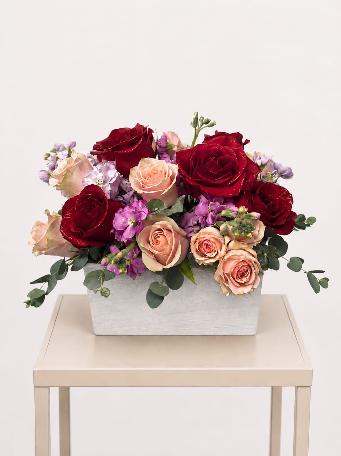 Red and blush roses with purple blooms in a white rectangular vase