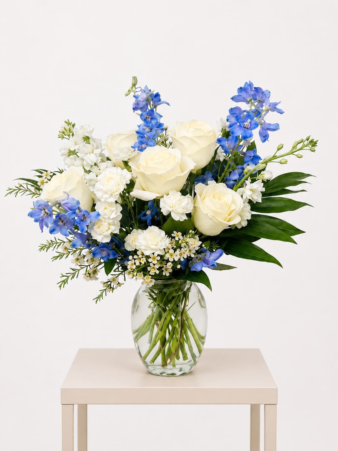 Elegant Blue &amp; Ivory Garden Bouquet - A refined hand-designed flower arrangement featuring premium ivory roses accented with soft blue delphinium, white carnations, delicate waxflower, and lush seasonal greenery. Beautifully arranged in a clear glass vase, this design blends freshness, elegance, and natural movement—creating a timeless and graceful floral gift.  Perfect for birthdays, sympathy, thank you gestures, get well wishes, welcoming a new baby, thinking of you, Mother’s Day, or adding a refined touch to home décor.  Available for local flower delivery throughout Naples, Bonita Springs, Estero, Fort Myers and Fort Myers Beach.  Designer’s Choice: Each arrangement is uniquely designed using the freshest seasonal flowers available. Flower varieties, colors and containers may vary while maintaining the overall style, value and beauty of the design.